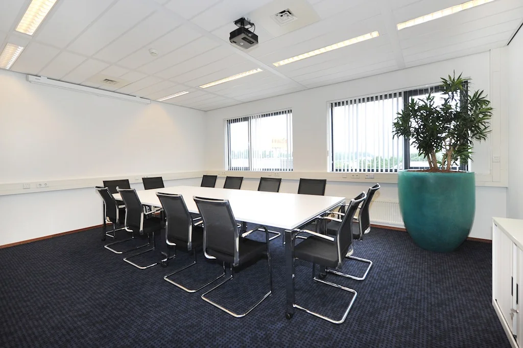 Vergaderruimte aan de Laan van s-Gravenmade met moderne inrichting, vergadertafel, stoelen, grote plant en ramen met verticale jaloezieën.